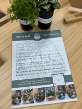 Load image into Gallery viewer, Instructions for building a terrarium on a wooden surface with potted plants.
