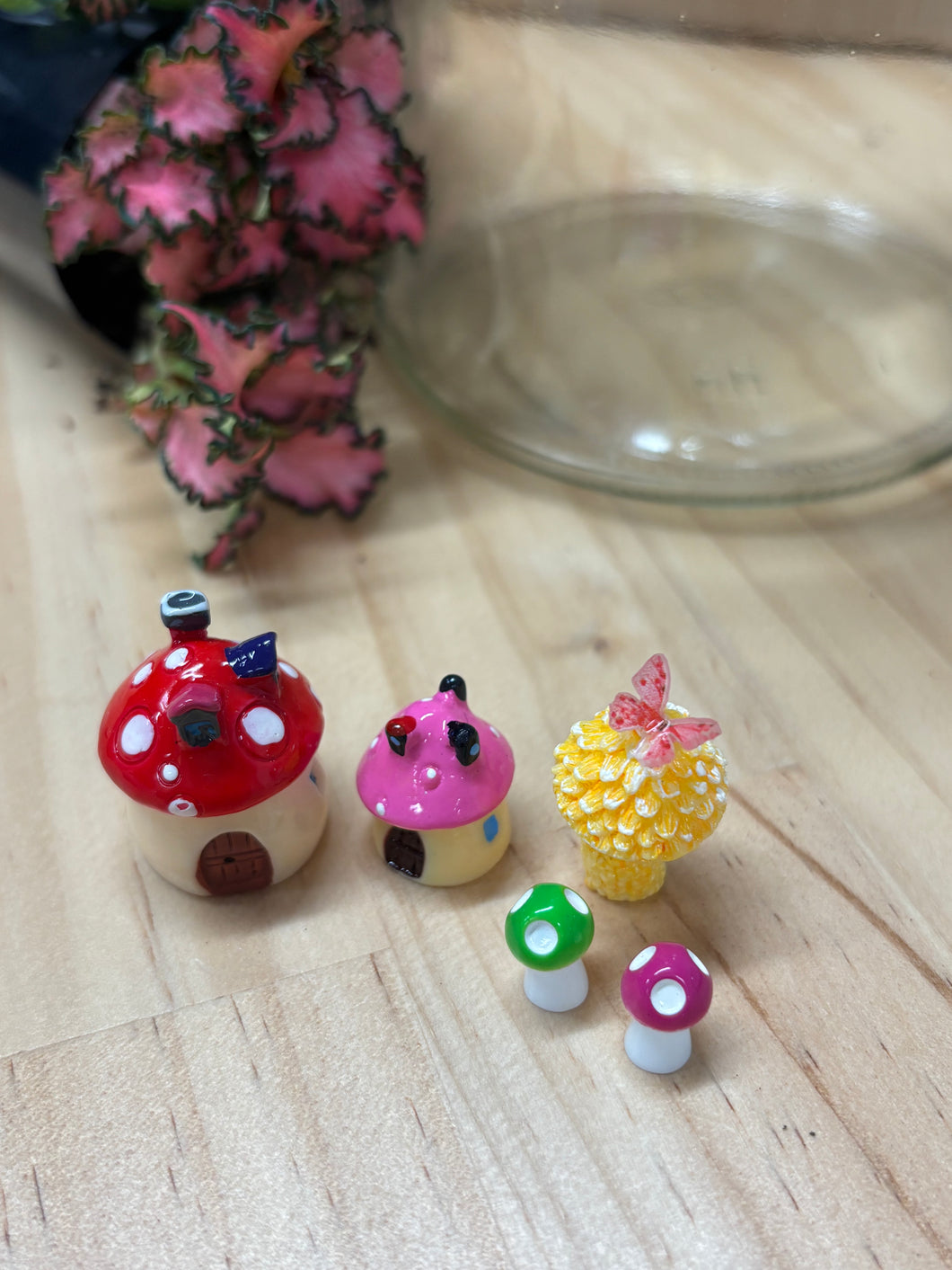 Mini fairy garden figurines sitting on a timber bench with glass jar and mini plants in the background.